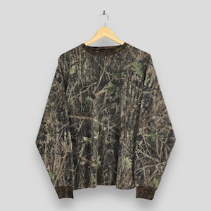 May include: Long-sleeved camouflage shirt with a crew neck and brown cuffs. The shirt features a realistic forest pattern in shades of brown, green, and tan, mimicking tree bark and foliage. The shirt is hanging on a wooden hanger.