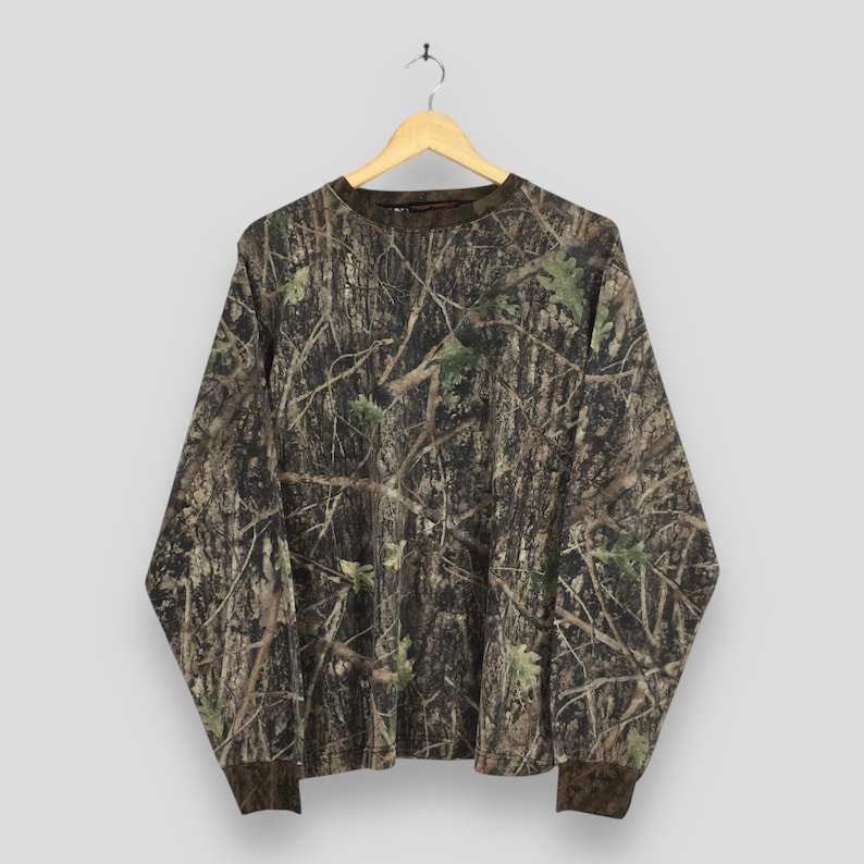 May include: Long-sleeved camouflage shirt with a crew neck and brown cuffs. The shirt features a realistic forest pattern in shades of brown, green, and tan, mimicking tree bark and foliage. The shirt is hanging on a wooden hanger.