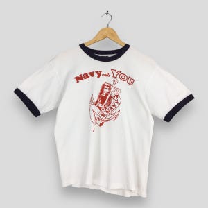 May include: White t-shirt with navy blue trim and sleeves, featuring a red graphic of a woman on an anchor with the text "Navy calls You" and "U.S. Navy". The shirt is hanging on a wooden hanger.