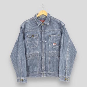 May include: A blue and white pinstriped denim jacket featuring a classic collar, a full-length front zip, two chest pockets with button flaps, and two lower slanted pockets. A small rectangular label with red text is sewn onto the left chest pocket.