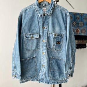 May include: A light blue denim chore coat featuring a pointed collar, a button-up front, and four front pockets. The jacket has long sleeves with buttoned cuffs and a small rectangular label on the upper left chest pocket with the text GL HEART.