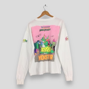 May include: White long-sleeve shirt with a colorful graphic on the back. The graphic features a green monster in front of a cityscape with the text "Yuppie Surf Street" and "Monstr". The sleeves have a small graphic.