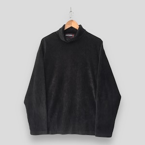 May include: A black long-sleeve turtleneck jumper featuring a vertical ribbed texture throughout the fabric. The garment includes a small rectangular label inside the collar with the text Lucky Strike in white letters on a red background.