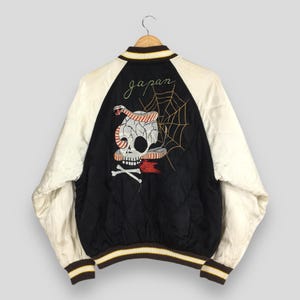 May include: A vintage black and white bomber jacket with a skull and snake embroidered design on the back. The jacket has white sleeves, a black quilted back, and the word "Japan" embroidered above the skull.