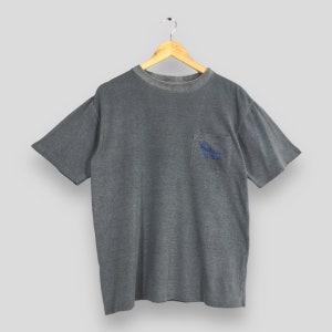May include: Gray short-sleeved t-shirt with a pocket on the chest. The pocket features blue text that reads "Autry U.S.A.". The shirt is hanging on a wooden hanger against a light gray background. The shirt has a slightly faded appearance.