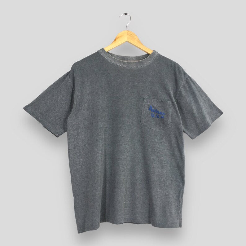 May include: Gray short-sleeved t-shirt with a pocket on the chest. The pocket features blue text that reads "Autry U.S.A.". The shirt is hanging on a wooden hanger against a light gray background. The shirt has a slightly faded appearance.