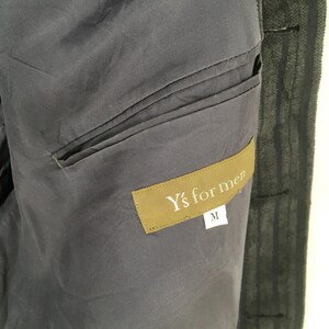 May include: A dark gray jacket interior featuring a horizontal welt pocket and a rectangular brown fabric label stitched onto the lining. The label displays the white text Y's for men with a smaller white tag below it showing the size letter M.