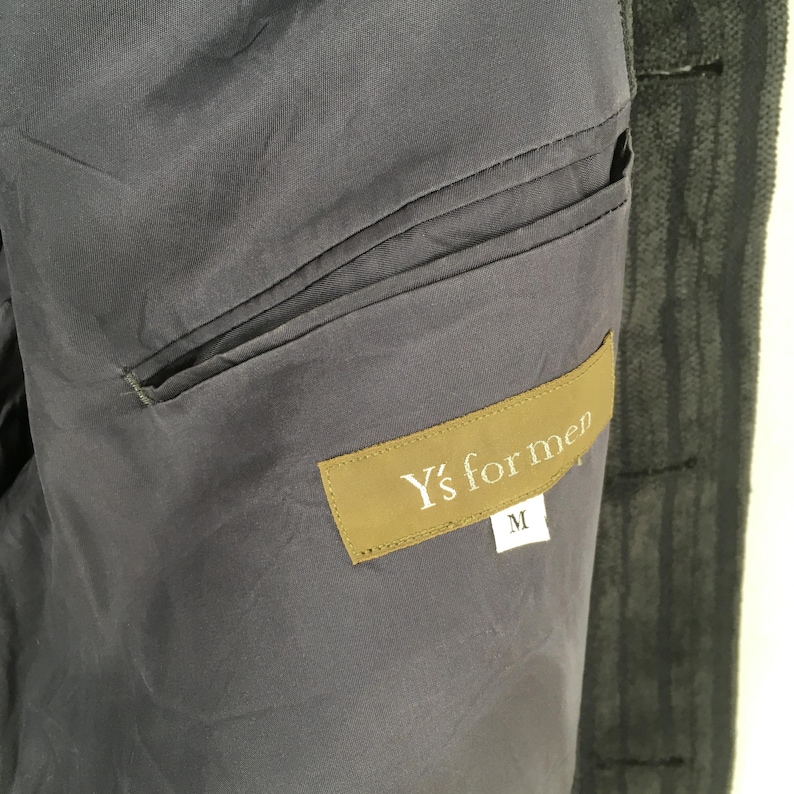 May include: A dark gray jacket interior featuring a horizontal welt pocket and a rectangular brown fabric label stitched onto the lining. The label displays the white text Y's for men with a smaller white tag below it showing the size letter M.