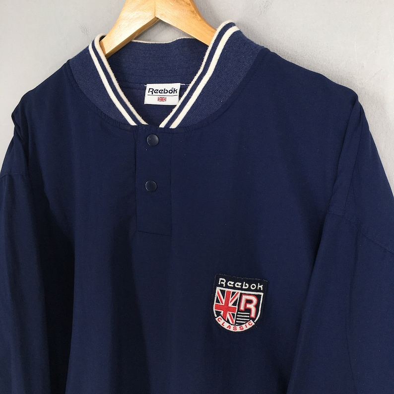May include: A navy blue long-sleeve shirt with a white and navy blue ribbed collar. The shirt has a button placket and a Reebok Classic logo patch on the left chest.