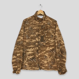 Puede incluir: Una chaqueta de camuflaje marrón con cierre de cremallera y cuello alto. La chaqueta tiene un estampado de palmeras y un pequeño bolsillo en el pecho izquierdo.