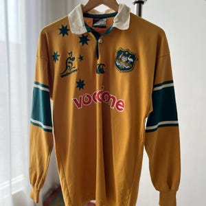 May include: A gold long-sleeve rugby shirt with a white collar, featuring a green Wallabies logo, dark green sleeve bands with white stripes, and the text Vodafone in red. The shirt includes a crest with an emu and kangaroo on the chest.
