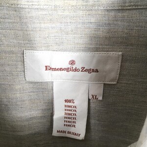 May include: Close-up of a light gray shirt with a label that reads "Ermenegildo Zegna." The label also states "100% Tencel" and "Made in Italy." The shirt is size XL.