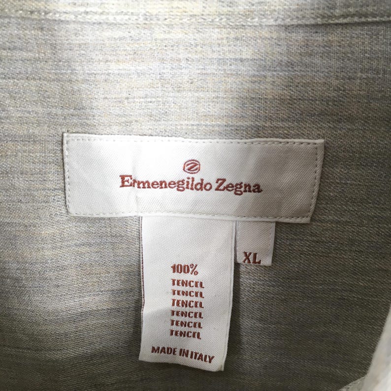 May include: Close-up of a light gray shirt with a label that reads "Ermenegildo Zegna." The label also states "100% Tencel" and "Made in Italy." The shirt is size XL.