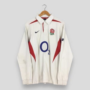 May include: A white long-sleeve rugby shirt featuring a collar, a button placket, and red side panels. The chest displays a black Nike swoosh logo on the right, a red and green rose emblem on the left, and a large blue O2 logo in the center.