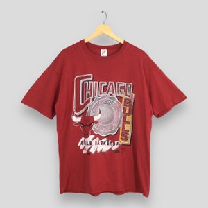 Puede incluir: Camiseta roja con un estampado gráfico del logotipo de los Chicago Bulls. El logotipo presenta un toro rojo con cuernos negros y un balón de baloncesto. El texto "Chicago Bulls" está impreso en letras blancas y amarillas.