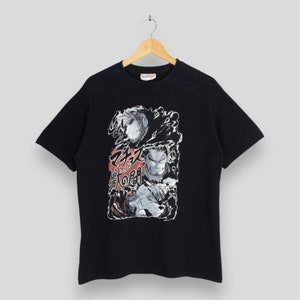 Puede incluir: Camiseta negra con un diseño gráfico que presenta dos personajes ilustrados y texto japonés. El diseño utiliza tonos grises, rojos y blancos. La camiseta está colgada de una percha de madera.
