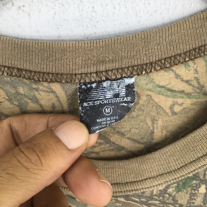 Puede incluir: Primer plano de una camiseta con estampado de camuflaje con cuello y ribetes de manga marrones. La etiqueta dice "ACE SPORTSWEAR" con una talla "M" y "MADE IN U.S.A."