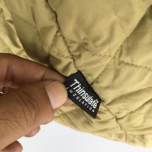 May include: Close-up of a tan quilted fabric with a black label that reads "Thinsulate INSULATION". The fabric has a diamond pattern. The label is being held by a hand.