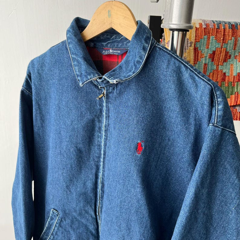 May include: A blue denim jacket with a full-length front zipper, a pointed collar, and long sleeves. The interior features a red and black plaid lining. A small red embroidered logo of a person on a horse is visible on the left chest area.