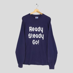 May include: Navy blue long-sleeve shirt with the text "Ready Steady Go!" in white block letters. The shirt is hanging on a wooden hanger against a light gray background.
