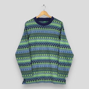 May include: A long-sleeved sweater with a crew neck, featuring a geometric pattern in shades of green, blue, and navy. The design includes repeating shapes like diamonds, triangles, and X's. The sweater is hanging on a wooden hanger.