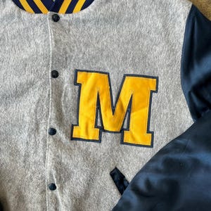 May include: Gray varsity jacket with a large yellow "M" patch. The jacket features navy blue satin sleeves and a navy blue collar with yellow stripes. The jacket has black buttons and a small pocket.