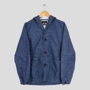 May include: A blue denim jacket with a hood, button closure, and two front pockets. The jacket is hanging on a wooden hanger. The jacket has a collar and a button closure. The jacket is made of a textured fabric.