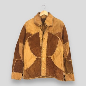 May include: A long-sleeve jacket constructed from patchwork panels of tan and dark brown suede leather. The garment features a pointed collar, a front button closure with four visible light-coloured buttons, and decorative whipstitch detailing.