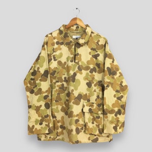 May include: A beige, brown and green camouflage print long-sleeved shirt with a zipped collar and a single patch pocket on the left side. The shirt is hanging on a wooden hanger.