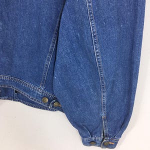 May include: Close-up of a blue denim jacket. The jacket features brass-colored buttons and seams. The sleeve is gathered at the wrist. The denim has a slightly faded appearance.