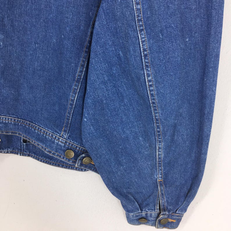 May include: Close-up of a blue denim jacket. The jacket features brass-colored buttons and seams. The sleeve is gathered at the wrist. The denim has a slightly faded appearance.