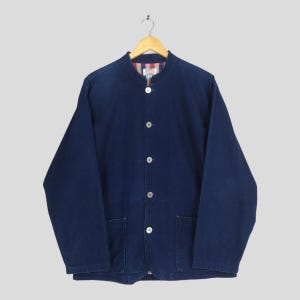 May include: A dark blue denim jacket with a mandarin collar and five white buttons. The jacket has two front pockets and a red and white striped lining visible at the collar. The jacket is hanging on a wooden hanger.