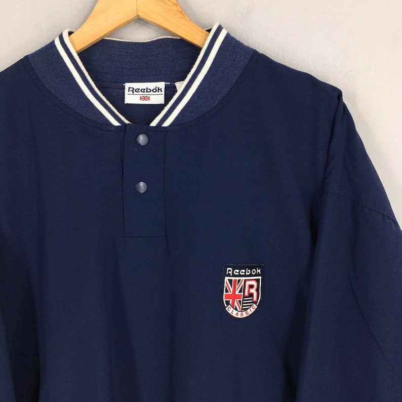 May include: A navy blue long-sleeve shirt with a white ribbed collar and a button-up placket. The shirt features a red and white embroidered Reebok logo with the text "Classic" underneath.