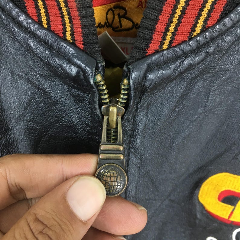 May include: Close-up of a black leather jacket with a brass zip. The jacket has a red, yellow, and black striped collar. The zip pull features a globe design. The jacket appears to be vintage.