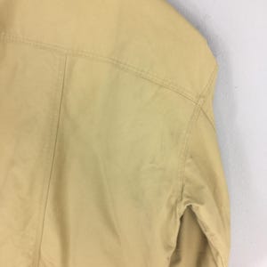 May include: Close-up of a tan jacket, showcasing the shoulder and upper back details. The jacket has visible stitching along the seams and a classic design. The fabric appears smooth and slightly shiny.
