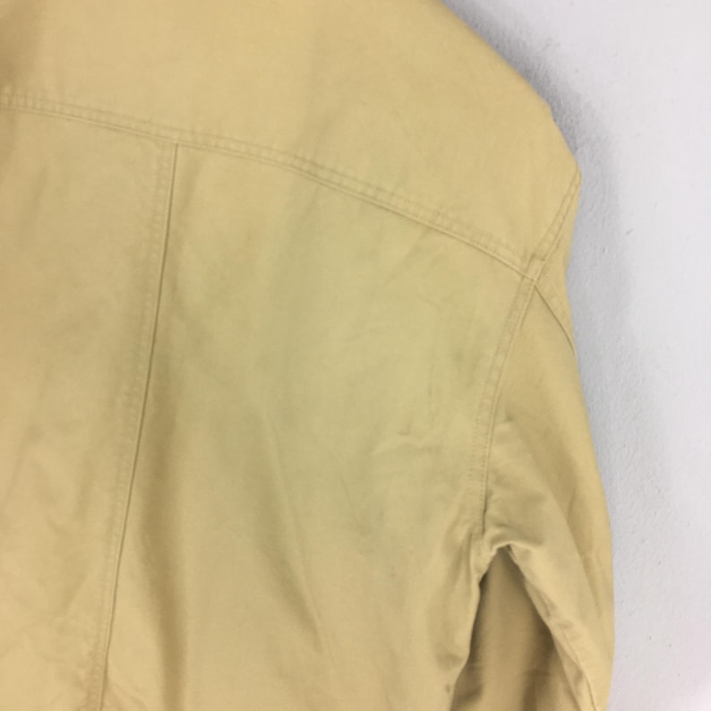 May include: Close-up of a tan jacket, showcasing the shoulder and upper back details. The jacket has visible stitching along the seams and a classic design. The fabric appears smooth and slightly shiny.
