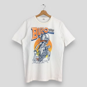 May include: White t-shirt with a cartoon graphic of a skeleton riding a motorcycle. The graphic includes the word "BUCO" in orange and blue, a checkered flag, and the word "PRODUCTS". The design is set against a yellow and orange background.
