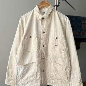 May include: An off-white Lee chore jacket featuring a pointed collar, a button-front fastening with metallic buttons, two chest pockets with button flaps, and two large lower patch pockets. A small Lee brand label is visible on the left chest pocket.