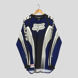 Op de afbeelding: Blauw, zwart en grijs shirt met lange mouwen en het Fox Racing-logo op de borst. Het shirt heeft witte accenten en het woord "RACING" verticaal op de mouwen gedrukt. Het hangt aan een houten hanger.