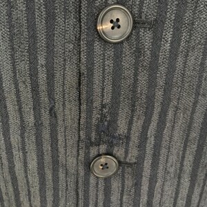 May include: A dark charcoal gray fabric garment featuring a vertical pinstripe pattern and two circular, four-hole buttons aligned vertically. The material has a textured, woven appearance with a visible seam running between the buttons.