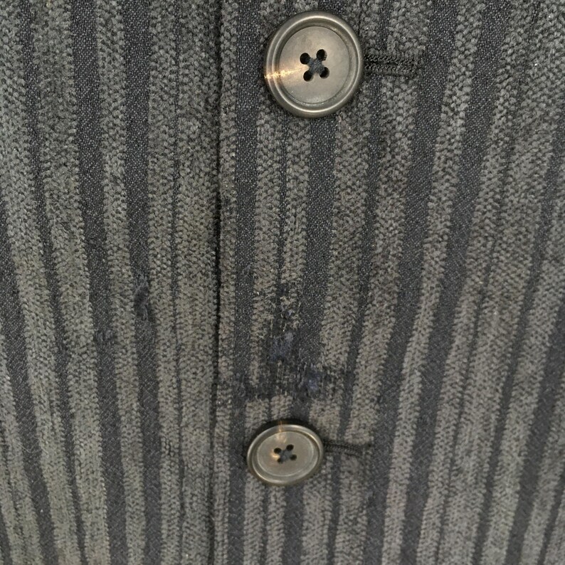 May include: A dark charcoal gray fabric garment featuring a vertical pinstripe pattern and two circular, four-hole buttons aligned vertically. The material has a textured, woven appearance with a visible seam running between the buttons.