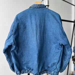 May include: A medium-wash blue denim jacket featuring a classic fold-down collar, long sleeves, and a gathered elasticized hemline. The jacket is displayed from the back, showcasing the solid denim fabric construction and the structured shoulder seams.