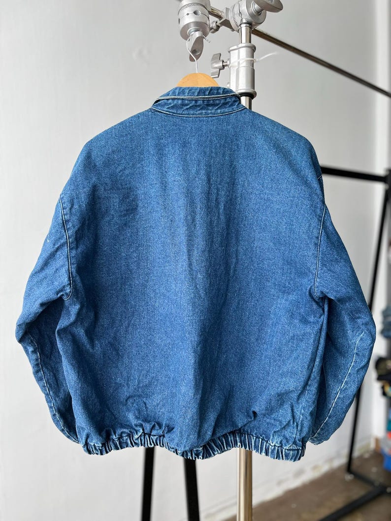 May include: A medium-wash blue denim jacket featuring a classic fold-down collar, long sleeves, and a gathered elasticized hemline. The jacket is displayed from the back, showcasing the solid denim fabric construction and the structured shoulder seams.