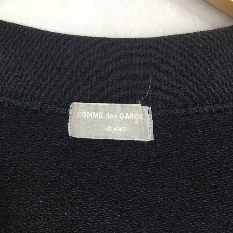 Puede incluir: Una etiqueta blanca con texto negro que dice "COMME des GARCONS" y "HOMME" sobre un fondo de tela negro.