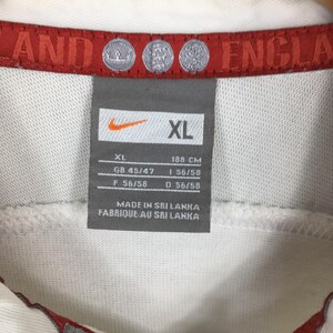 Peut inclure: Gros plan sur une &eacute;tiquette de chemise Nike blanche. L'&eacute;tiquette est grise avec le logo Nike et la taille XL. L'&eacute;tiquette comprend &eacute;galement des mesures en centim&egrave;tres et le texte "MADE IN SRI LANKA". La chemise a une bordure rouge.