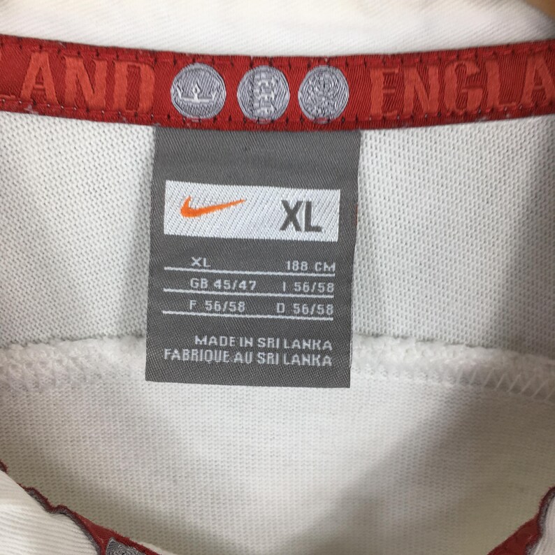 Peut inclure: Gros plan sur une &eacute;tiquette de chemise Nike blanche. L'&eacute;tiquette est grise avec le logo Nike et la taille XL. L'&eacute;tiquette comprend &eacute;galement des mesures en centim&egrave;tres et le texte "MADE IN SRI LANKA". La chemise a une bordure rouge.