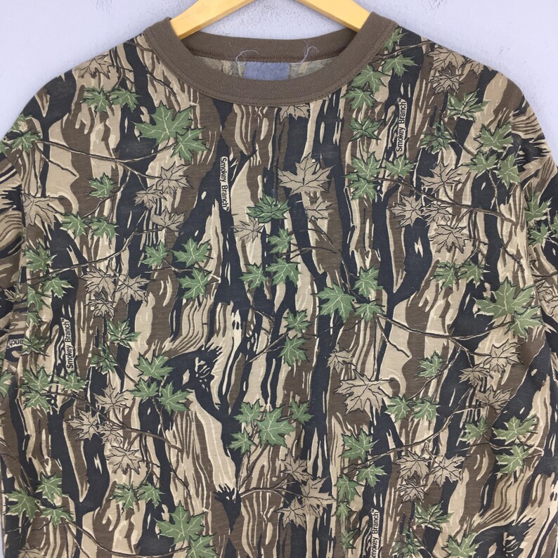 May include: A long-sleeved, crew neck shirt with a camouflage pattern. The shirt features a brown collar and a repeating pattern of green leaves and branches over a brown and black background. The text "Smokey Branch" is visible on the shirt.