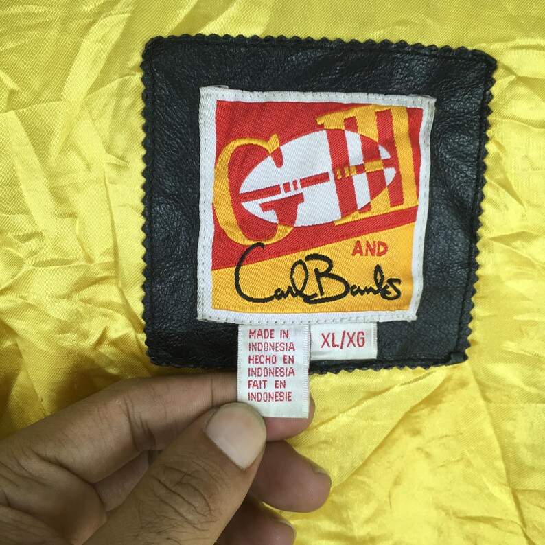 May include: A black leather label with the text "G and Carl Banks" in a red and white design. The label also includes text indicating the item was made in Indonesia. The background is yellow.