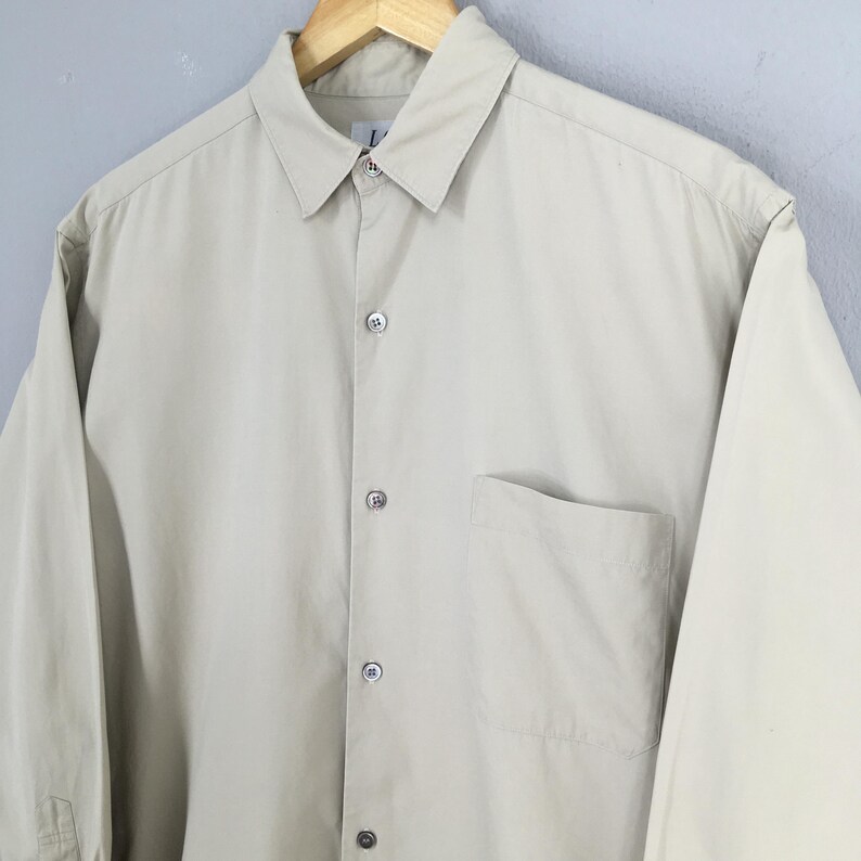 Peut inclure: Une chemise boutonn&eacute;e beige clair avec un col et une poche poitrine unique. La chemise &agrave; manches longues est faite d'un tissu lisse et boutonn&eacute;e. La chemise est suspendue &agrave; un cintre en bois.