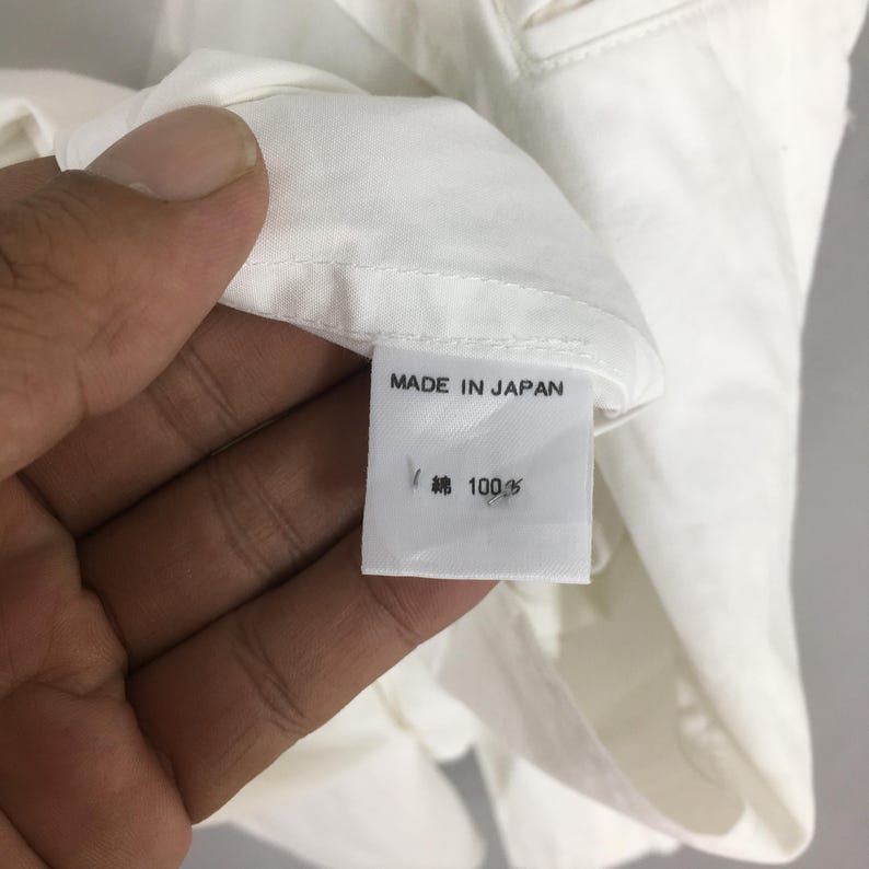 May include: A white garment featuring a rectangular fabric care label sewn into the interior seam. The label displays the text MADE IN JAPAN in black capital letters, followed by the Japanese characters for cotton and the number 100%.